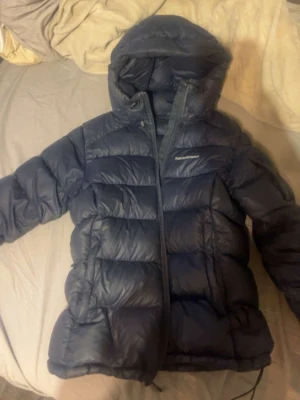 Mörkblå dunjacka från Peak Performance - Säljer en mörkblå dunjacka från Peak Performance med huva och dragkedja. Jackan har en puffig design med breda kanaler och logga på bröstet. Perfekt för kalla vinterdagar, säljer den för kommer inte till någon nytta då jag har andra, den ligger på 600 kr men kom annars med pris om de inte skulle passa 