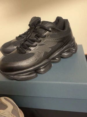 Svarta sneakers med futuristisk sula - Snygga helsvarta sneakers med mesh och syntetmaterial, samt en unik, chunky sula i flera lager för en futuristisk vibe. Skorna har snörning och sportig design som sticker ut. Perfekta för dig som gillar streetwear och vill ha något som syns.