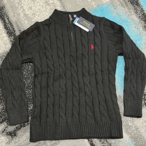 Svart Zip Knit Polo Ralph Lauren - Kabelstickad svart Zip Knit från Polo Ralph Lauren med halv dragkedja och röd broderad logga på bröstet. Tröjan har ribbade muddar och hög krage, perfekt för en stilren och klassisk look.TVEKA INTE ATT SKICKA EN BUD:)