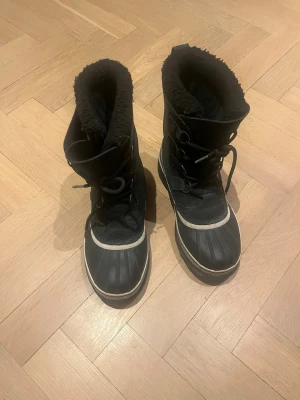 Svarta vinterstövlar från Sorel - Rejäla svarta vinterstövlar från Sorel med snörning och grov sula. Insidan är fodrad med mjukt, svart material för extra värme. Yttermaterialet är slitstarkt och vattenavvisande, perfekt för kalla dagar. Klassisk design med vit rand runt sulan.