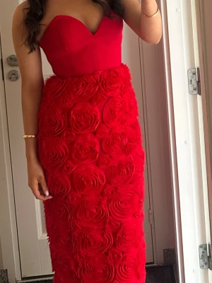 Röd långklänning med rosdetaljer - Säljer en stunning röd långklänning med hjärtformad urringning och axelbandslös design. Kjolen är täckt av stora 3D-rosor som ger en unik och lyxig känsla. Perfekt för dig som vill sticka ut på bal eller fest! 💃sluta där bak, pris går att diskutera. Eftersom det är snörning där bak är inte storleken till bara just 36 utan kan funka andra med inte så stor storleks skillnad.🩷