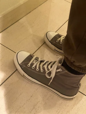 Gråa Converse Chuck Taylor All Star - Säljer ett par klassiska höga Converse Chuck Taylor All Star i grå canvas med vita gummisulor och tåhättor. Skorna har vita snören och metallöljetter, samt den ikoniska loggan på sidan. Perfekta för en avslappnad och stilren look. Storlek 44,5