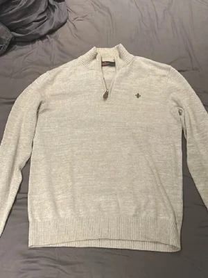 Beige stickad tröja med halvzip - Snygg beige stickad tröja från Morris med halvzip och ribbade muddar. Tröjan har en liten broderad logga på bröstet och hög krage. Perfekt för lager på lager och enkel att matcha med olika outfits.