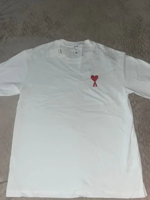 Vit t-shirt från Ami med rött hjärta - Vit t-shirt från Ami med klassisk passform och rund hals. På bröstet finns ett broderat rött hjärta med bokstaven A. Tillverkad i mjuk bomull som känns skön mot huden. Perfekt för en clean och stilren look.