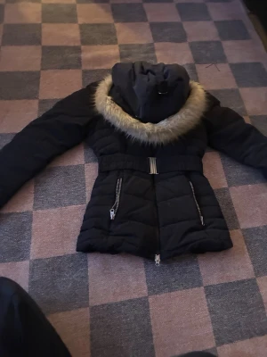 Mörkblå pufferjacka med fuskpäls - Snygg mörkblå pufferjacka med avtagbar huva och fuskpäls runt kragen. Jackan har två dragkedjeförsedda fickor framtill, markerad midja med bälte och quiltad design. Perfekt för kalla vinterdagar och riktigt varm. Har köpt för 4 tusen men säljer för 800 jag har it använt så mycket och den e som ny