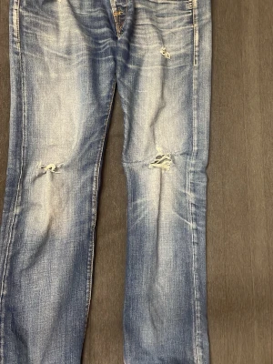 Blå slitna jeans med hål - Säljer ett par blå jeans med slitningar och hål på knäna. Jeansen har en tvättad look med ljusare partier och raka ben. Perfekta för en avslappnad och trendig stil. Materialet är klassisk denim i bomull.