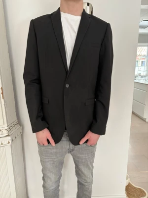 Svart klassisk kavaj herr - Stilren svart kavaj med smal passform och enkel knappstängning. Kavajen har två fickor med lock och en bröstficka. Perfekt till skjorta eller t-shirt för en clean look. Materialet ser ut att vara en blandning av bomull och polyester.  Det står XL men sitter som en L modellen är 186 cm lång och väger 75 kg och den sitter bra på han. Ny pris på en sån här är runt 10000 tusen!