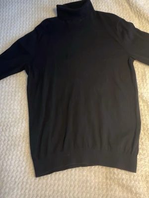 Svart polotröja stickad H&M - Klassisk svart stickad polotröja med lång ärm och ribbade muddar. Tröjan har en hög polokrage och är tillverkad i ett mjukt, tunt material som passar perfekt till lager på lager. Enkel och stilren design utan synliga loggor eller mönster.