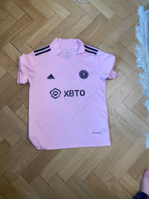 Inter Miami rosa matchtröja Adidas - Snygg rosa matchtröja från Inter Miami CF, tillverkad av Adidas med svart logga och klubbmärke på bröstet. Tröjan har svarta ränder på axlarna och är gjord i ett lätt och ventilerande material med AEROREADY-teknologi. Perfekt för fotbollsträning eller som supporterplagg. Och har en litet smuts i minnen