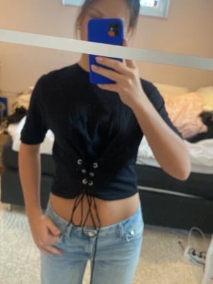 Svart croppad topp med snörning - Säljer en svart croppad topp med snörning framtill. Toppen har korta ärmar och en tight passform som framhäver midjan. Perfekt för dig som gillar en edgy och trendig stil.