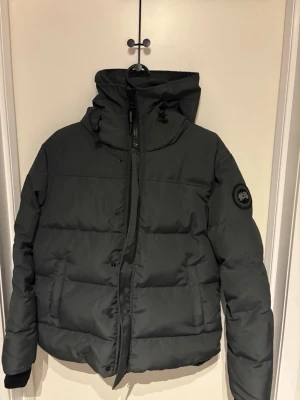 Svart pufferjacka från Canada Goose - Säljer en svart pufferjacka från Canada Goose med hög krage, huva och diskret logotyp på ärmen. Jackan har dragkedja och knappar framtill samt två stora fickor. Perfekt för kalla vinterdagar och riktigt snygg streetstil.