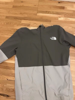 Grå vindjacka från The North Face - Snygg tvåfärgad vindjacka från The North Face i mörkgrått och ljusgrått. Jackan har hel dragkedja, huva och logga på bröstet. Tillverkad i ett lätt och vindtätt material, perfekt för blåsiga dagar. Storlek S