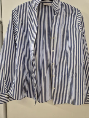 Skjorta - Klassisk skjorta från Chicos i blåvitt randigt mönster. Skjortan har lång ärm, knappar framtill och en tidlös krage. Tillverkad i bomull för en skön och luftig känsla. Perfekt för dig som gillar stilrena och fräscha looks.