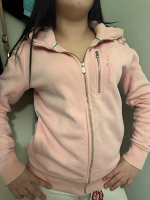 Ljusrosa hoodie från Sail Racing - Ljusrosa hoodie från Sail Racing med dragkedja framtill och en extra dragkedjeförsedd bröstficka. Hoodien har ribbade muddar vid ärmslut och nederkant samt broderad logga på bröstet. Perfekt för en avslappnad och sportig stil.