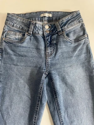 Blå raka jeans från Lemon Lush - Snygga blå jeans från Lemon Lush med lite utsvängar och klassisk femficksdesign. Jeansen har dubbla knappar i midjan och ljus tvätt för en avslappnad look. Tillverkade i mjukt denimtyg som är skönt att bära hela dagen.