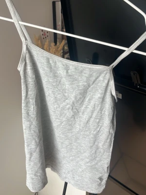 Ljusgrått linne med smala axelband - Snyggt ljusgrått linne med smala axelband och rak passform. Tillverkat i mjukt och stretchigt material som känns skönt mot huden. Perfekt att styla lager på lager eller bära som det är under varma dagar.