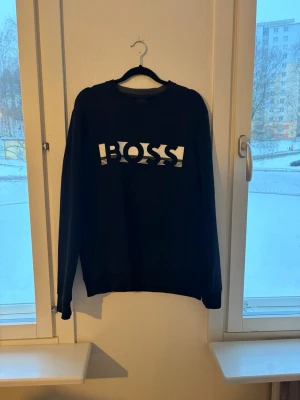 Svart sweatshirt från BOSS - Säljer en svart sweatshirt från BOSS med stor vit logga framtill. Tröjan har rund halsringning och långa ärmar. Tillverkad i mjukt material som passar perfekt till en avslappnad stil. Enkel och stilren design med ribbade muddar.