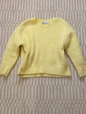 Gul stickad tröja från Zara - Säljer en ljusgul stickad tröja från Zara med rund hals och ribbstickade detaljer. Tröjan har en avslappnad passform och långa ärmar, perfekt för att hålla sig varm under kyliga dagar. Materialet är mjukt och fluffigt vilket gör den extra bekväm.