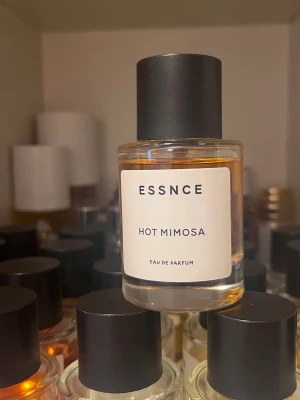 ESSNCE Hot Mimosa Eau de Parfum - ESSNCE Hot Mimosa Eau de Parfum kommer i en stilren, rund glasflaska med svart lock och minimalistisk vit etikett. Doften har en varm, gyllene färg som ger ett lyxigt intryck. Perfekt för dig som gillar moderna och eleganta parfymer.
