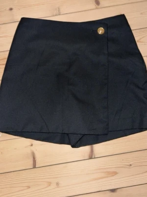 Svart skort med guldfärgad knapp - Stilren svart skort med omlott-look och en snygg guldfärgad knapp i midjan. Klassisk rak passform och diskret shortsdel undertill. Perfekt för dig som vill ha kjol-looken men ändå känna dig bekväm. Materialet känns mjukt och följsamt.