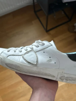 Vita sneakers från Philippe Model - Stilrena vita sneakers från Philippe Model med klassisk låg profil och svarta detaljer vid hälen. Skorna har vita skosnören, diskret logga på sidan och är tillverkade i läder. Perfekta för en clean och avslappnad look.