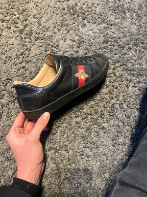 Gucci Ace svarta sneakers med bi - Gucci Ace sneakers i svart läder med röd och grön sidoremsa och broderad guldfärgad bi. Skon har svarta skosnören, mörkblå detalj vid hälen med Gucci-logga och beige insida. Klassisk låg modell med exklusiv känsla och ikoniska detaljer. Utan box