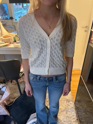 Vit kortärmad kofta från Kappahl - Söt vit kofta från Kappahl med korta ärmar och v-ringning. Koftan har ett fint hålmönster, ribbade muddar och knappar framtill. Perfekt att slänga över en topp för en fräsch look. Materialet känns mjukt och luftigt.