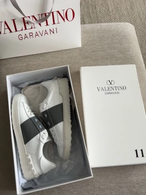 Valentino open grå - Snygga vita sneakers från Valentino med bred grå rem över mitten och nitar längs hälen. Skorna har en låg profil, vita snören och en chunky sula. Tillverkade i slätt läder för en clean och modern look. Storlek är 39-40. Skriv om mer bilder!