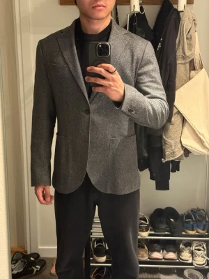 Riley kavaj - Snygg grå kavaj från Riley med två knappar och klassisk passform. Kavajen har smala slag, två framfickor och är tillverkad i en mjuk ullblandning. Perfekt för dig som vill ha en stilren och tidlös look. Kavajen är i utmärkt skick utan skavanker och defekter. Modellen är ca 175 