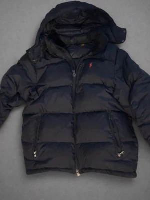 Mörkblå pufferjacka från Polo Ralph Lauren - OBS Storlek L men passar M!                         Snygg mörkblå pufferjacka från Polo Ralph Lauren med röd logga på bröstet. Jackan har avtagbar huva, två dragkedjefickor och kraftig vaddering som håller dig varm. Perfekt för kalla vinterdagar och har en klassisk, sportig look.
