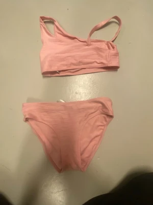 Ljusrosa bikini med asymmetrisk topp - Snygg ljusrosa bikini med sportig, asymmetrisk topp och matchande trosor. Bikinin har enkel design utan mönster och smala axelband. Perfekt för strandhäng eller poolen.