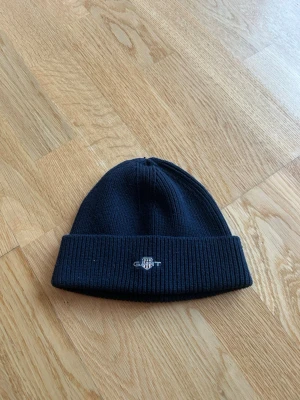 Mörkblå stickad mössa från GANT - Snygg mörkblå stickad mössa från GANT med broderad logga framtill. Mössan har uppvikt kant och är tillverkad i mjukt material som håller dig varm. Perfekt för kyliga dagar och enkel att matcha med din jacka.