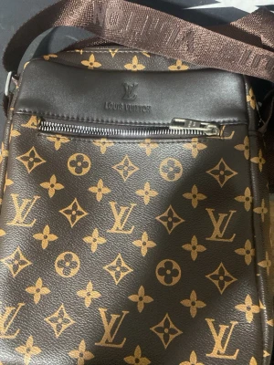 Louis Vuitton brun axelväska - Snygg brun axelväska från Louis Vuitton med klassiskt monogrammönster i beige och guldtoner. Väskan har justerbar axelrem, dragkedja fram och bak samt detaljer i konstläder. Perfekt för dig som gillar lyxig och tidlös design. (Lappen är lite sliten men sitter kvar)
