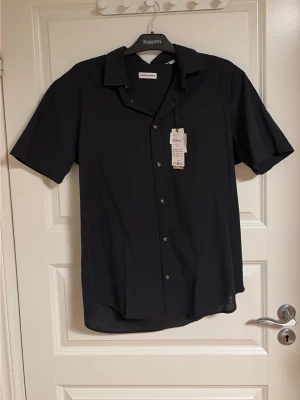 Svart kortärmad skjorta  - Stilren svart skjorta från Jack & Jones med korta ärmar och klassisk krage. Skjortan har knappar framtill och en avslappnad passform, perfekt för en enkel och snygg look. Tillverkad i mjukt material som känns skönt mot huden.