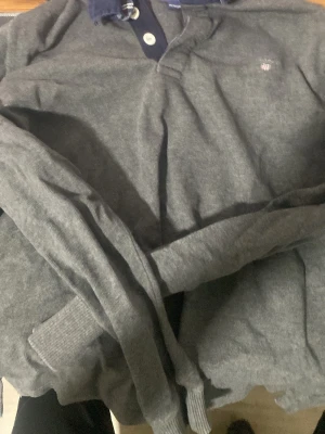 Grå långärmad tröja från Gant - Mysig grå långärmad tröja från Gant med mörkblå krage och knappslå. Tröjan har små vita knappar och en diskret Gant-logga broderad på bröstet. Perfekt till jeans eller chinos för en avslappnad men stilren look.        (PSST STORLEKEN PÅ ANNONSEN ÄR FEL, DEN RIKTIGA SYNS PÅ BILDEN OCH ÄR 11/12 ÅR OCH ÄR 146/152CM)