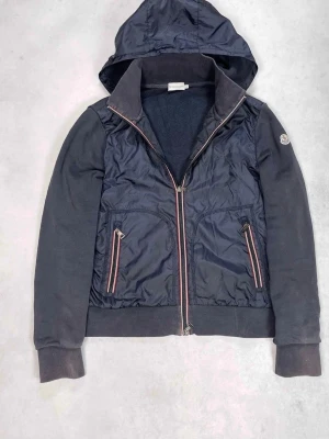 Moncler cardigan - Stilren svart cardiganjacka från Moncler med quiltad front och mjuka ärmar. Jackan har dragkedja framtill, två fickor med rödvita detaljer och Moncler-logga på ärmen. Perfekt för lager-på-lager och snygg streetstil. Möts i Stockholm 