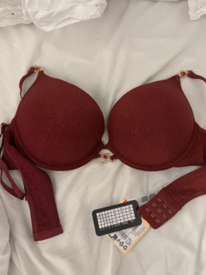 Vinröd push-up bh med glitterdetaljer - Snygg vinröd push-up bh från Lindex med glittrande material och dekorativa guldfärgade detaljer vid axelbanden och mellan kuporna. Bh:n har justerbara axelband och flera hakar för perfekt passform. Perfekt för dig som vill ha något extra.