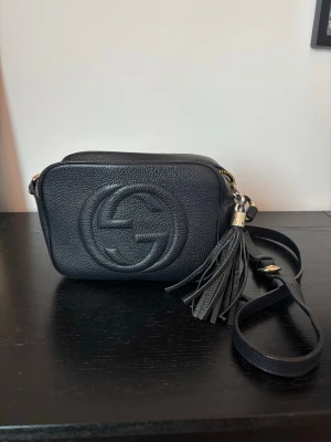 Svart axelväska modell Soho - Snygg svart axelväska (modell Soho). Väskan har justerbar axelrem och en hängande tofs vid dragkedjan. Perfekt storlek. Dragkedjan behöver bytas, litet hål inuti. Annars inga defekter.