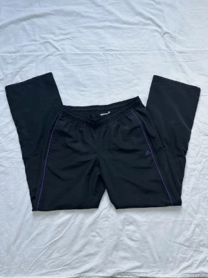 Adidas byxor  - Svarta Adidas byxor med lila sömmar längs benen och broderad logga på vänster sida. Byxorna har resår i midjan och raka ben. Perfekta för chill eller träning, tillverkade i ett lätt och mjukt material.