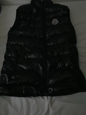 Svart dunväst från Moncler - Snygg svart dunväst från Moncler med glansig finish och klassisk logga på bröstet. Västen har hög krage och dragkedja framtill. Perfekt för lager-på-lager och ger en cool streetwear-look.