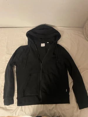 Svart hoodie med dragkedja Jack & Jones - Klassisk svart hoodie från Jack & Jones med dragkedja framtill, två fickor och justerbar huva med snören. Tillverkad i mjukt bomullsmaterial som är skönt mot huden. Perfekt för en avslappnad och stilren look.