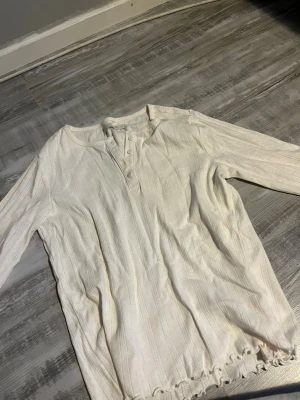Vit ribbad långärmad tröja 157 - Säljer en vit långärmad tröja från 157 med ribbad struktur och knappar vid halsen. Tröjan har en avslappnad passform och lätt vågig kant nedtill. Perfekt för en enkel och stilren look.