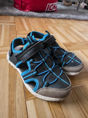 Svarta sportiga sandaler med blå detaljer - Säljer ett par svarta sportiga sandaler med blå detaljer och kardborreband. Sandalerna har en robust sula och öppet tåparti, perfekta för aktiva dagar. Materialet är en mix av syntet och textil med förstärkt tå. Snygg design med elastiska band för extra stöd.