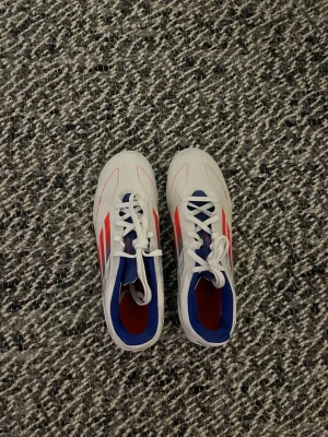 Vita Adidas F50 fotbollsskor - Säljer ett par vita Adidas F50 fotbollsskor med röda och blå detaljer. Skorna har klassisk snörning, låg profil och är tillverkade i syntetmaterial för lätt vikt och bra bollkontroll. Insidan är blå och yttersulan är designad för bra grepp på planen.
