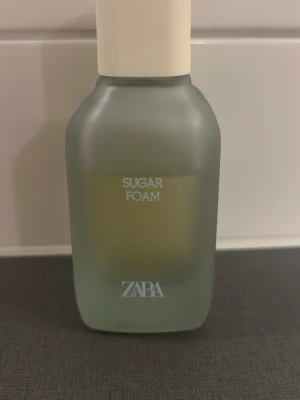 ZARA Sugar Foam parfym - ZARA Sugar Foam parfym i en stilren, frostad glasflaska med mjuka former och vit kork. Flaskan har en modern design och texten är tryckt i vitt. Perfekt för dig som gillar söta och fräscha dofter.