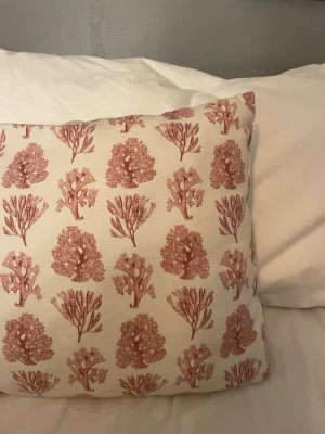 Beige kudde med röd korallmönster - Dekorativ kudde i beige tyg med tryckt röd korallmönster. Kudden har ett rektangulärt format och passar perfekt som prydnad på sängen eller soffan. Mönstret består av olika korallmotiv i en varm röd ton.