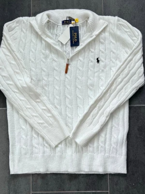 Vit Polo Ralph Lauren - Kabelstickad vit tröja från Polo Ralph Lauren med halv dragkedja och ribbade muddar. Klassisk broderad logga i svart på bröstet och mjukt material som känns lyxigt. Perfekt för dig som gillar stilrena och tidlösa plagg.