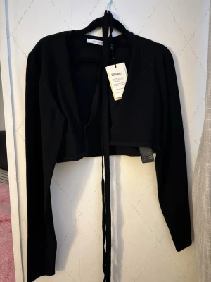 Svart cropped cardigan med knytband - Trendig svart cropped cardigan från EBIvario med långa ärmar och knytband framtill. Perfekt att styla över ett linne eller klänning för en cool lager-på-lager-look. Enkel och stilren design som passar till mycket.