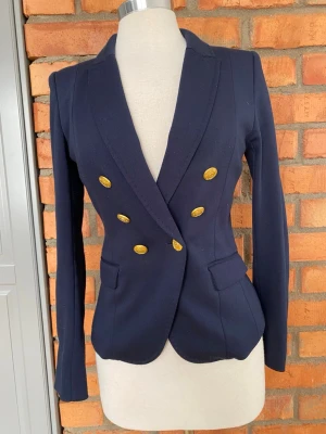 H&M Mörkblå kavaj med guldknappar strl 38 - Snygg mörkblå kavaj med dubbelknäppning och sex dekorativa guldknappar framtill. Kavajen har figurnära passform, klassisk krage och två fickor med lock. Långa ärmar med matchande guldknappar vid ärmslut. Perfekt för en stilren och trendig look. Helfordrad.