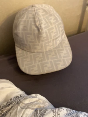 Beige Fendi keps med FF-mönster - Säljer en beige keps från Fendi med det klassiska FF-logomönstret över hela kepsen. Kepsen har böjd skärm och är tillverkad i ett tygmaterial som känns både lyxigt och stilrent. Perfekt för dig som vill ha en exklusiv accessoar till din outfit.